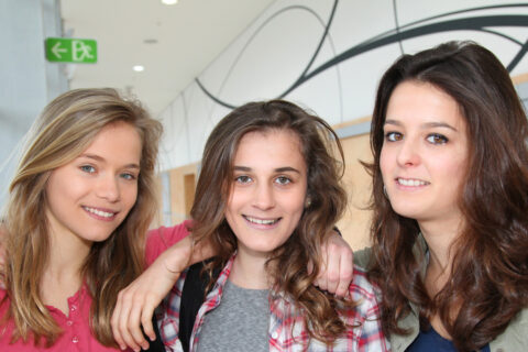 Drei Schülerinnen stehen in einem Gang einer Schuler und blicken in die Kamera