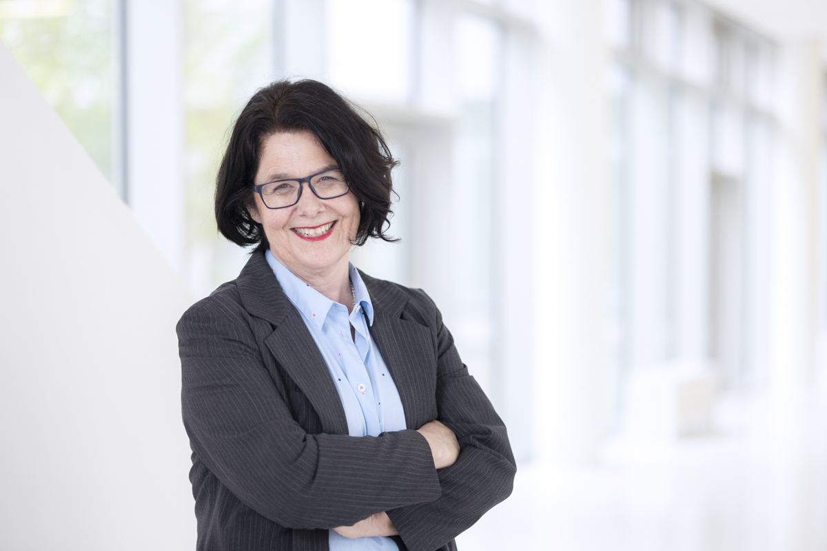 Eine Person mit dunkler Jacke über einer hellen Bluse steht mit verschränkten Armen vor einer weißen Wand mit Fenstern. Die Person hat schwarze Haare, trägt eine Brille und blickt lächelnd in die Kamera.