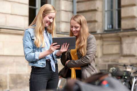 Zwei Studentinnen sehen sich etwas auf einem Tablet an, im Hintergrund ist ein Teil des Kollegienhauses zu sehen.