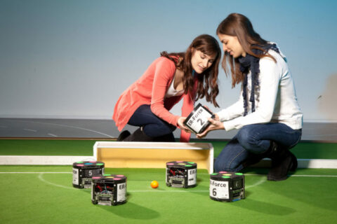 Zwei Frauen einen Spielroboter auf einem Spielfeld genauer an.