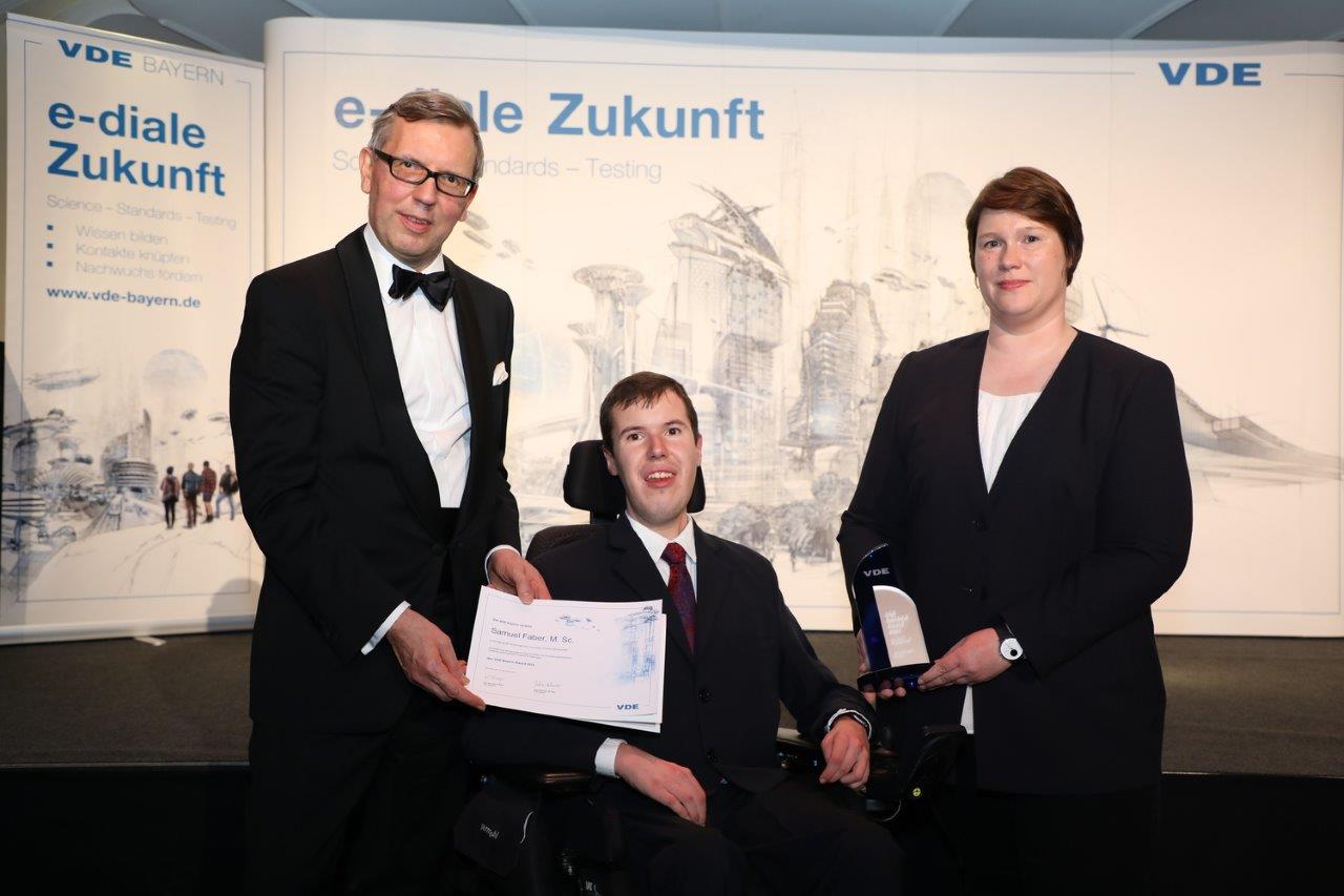 Vor einer Wand stehen 2 Personen und 1 Person im Rollstuhl, alle haben einen schwarzen Anzug an. Die Person im Rollstuhl hält eine Urkunde in der Hand.