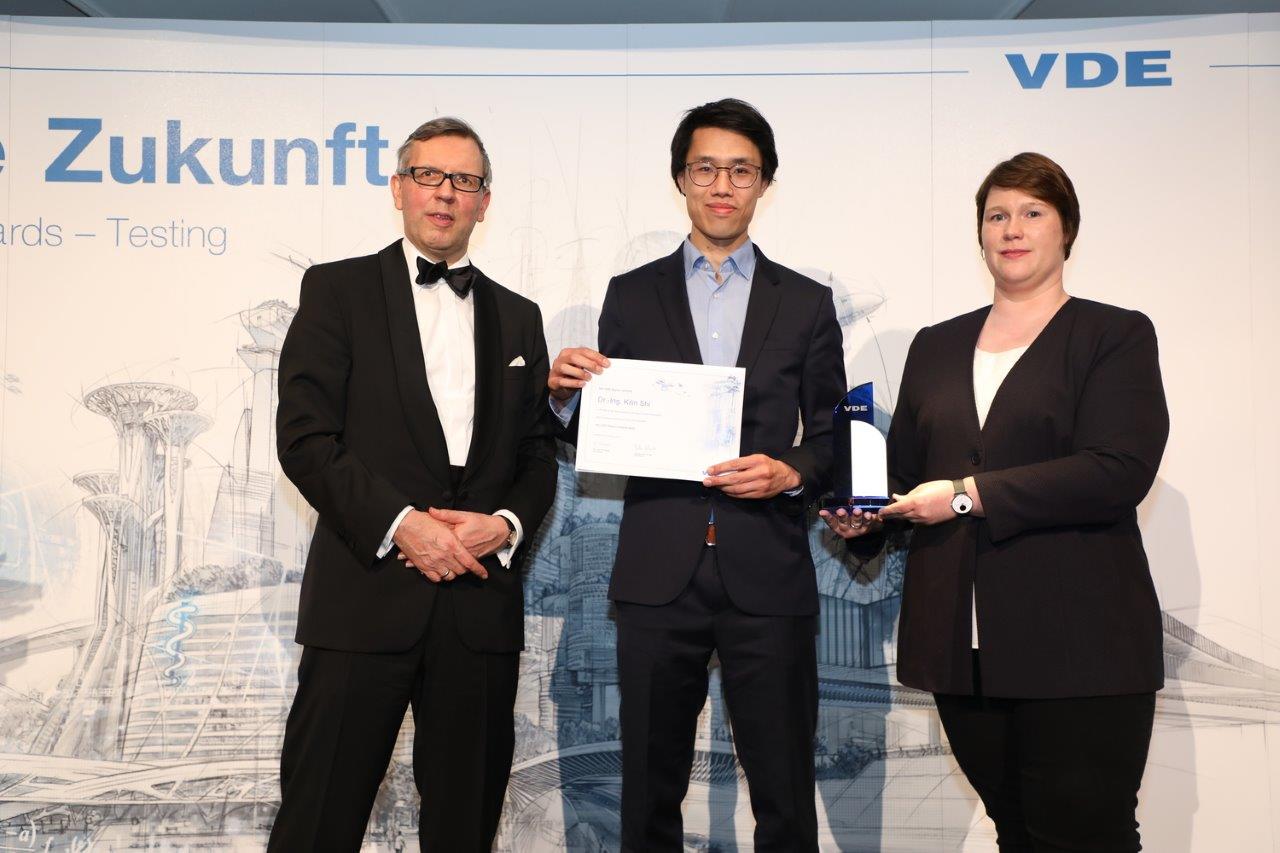 3 Personen in schwarzen Anzügen stehen vor einer Wand. Die Person in der Mitte hält eine Urkunde in der Hand.