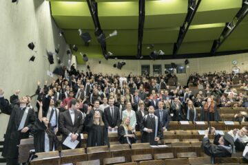 Viele Personen stehen beieinander und blicken in die Kamera. Einige von ihnen werfen ihre Absolventenhüte freudig in die Luft.