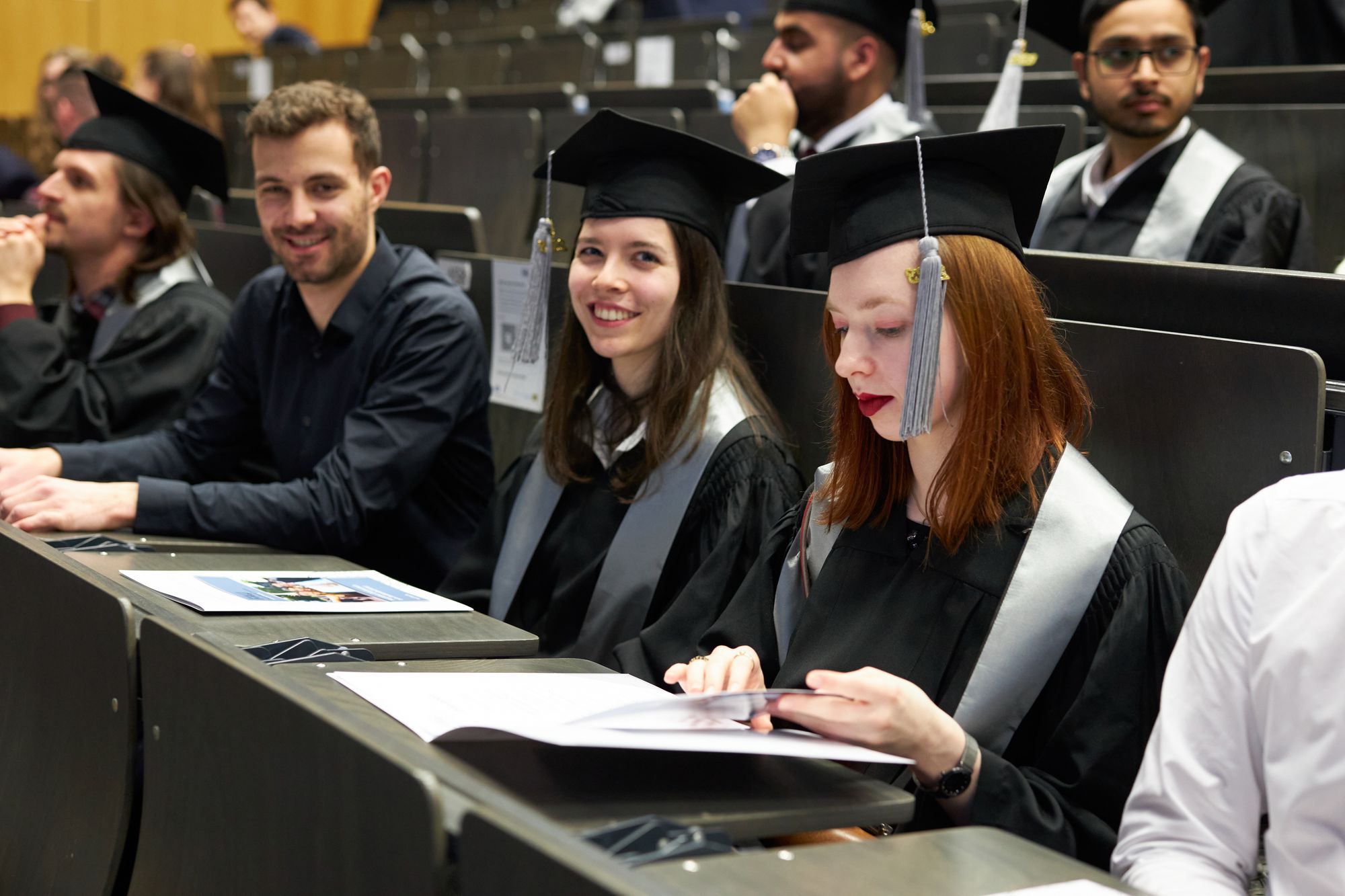 Mehrere Personen sitzen in einem Hörsaal, vor ihnen liegt die Absolventenurkunde.