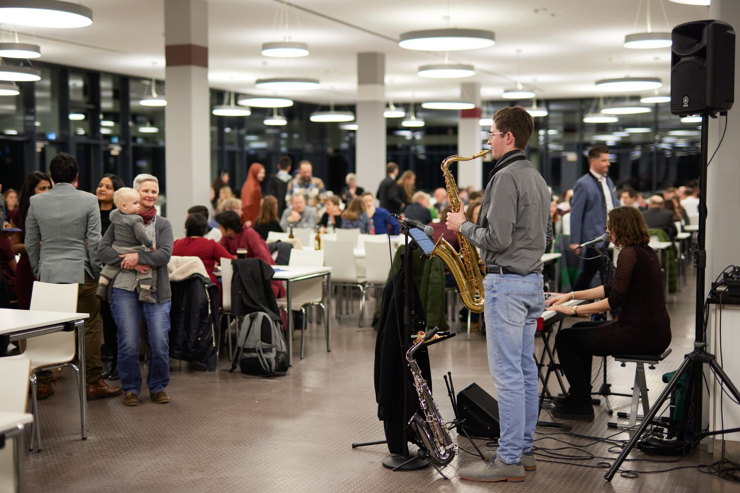 Ein Saxophonist und eine Klavierspielerin spielen Musik vor mehreren Personen.