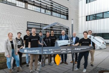 11 Personen stehen vor grauen Gebäude, sie halten eine große, schwarze Drohne in der Hand.
