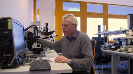 Ein Mann mit weißen Haaren und Brille sitzt an einem Schreibtisch und schaut in einen Bildschirm. Im Hintergrund sind gelbumrandete Fenster zu sehen.