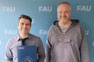 2 Personen stehen vor einer blauen Wand mit dem Logo der FAU und blicken lächelnd in die Kamera. Die eine Person hat eine blaue Urkundenmappe der FAU in der Hand.