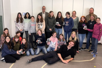 Viele junge Frauen sitzen und stehen beieinander, einige halten ein elektronisches Gerät in der Hand.