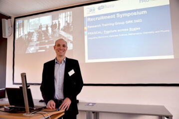 Dr. Sebastian Pfaller steht vor einer Leinwand, auf die eine Präsentationsfolie zu einem Symposium gezeigt wird.