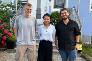 3 persons standing in front of a house. From left to right: Prof. Robert Schober, Dr. Gui Zhou, Dr. Vasileios Papanikolaou
