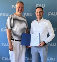 Zwei Männer in heller Kleidung stehen vor einer hellblauen Wand. Der Mann auf der rechten Seite hält eine geöffnete Urkunde