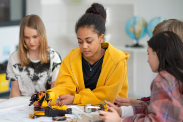 An einem Tischen sitzen vier Jugendliche, sie schauen sich bzw. fassen ein Gerät an.