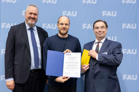 3 Personen stehen nebeneinander vor einer blauen Wand mit dem Schriftzug FAU. Die Person in der Mitte hält eine Urkunde in die Kamera, die Person daneben hält ein gelb eingepacktes Geschenk in der Hand.