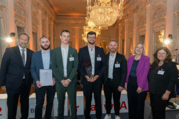 7 Personen stehen in einem festlichen Raum mit Kronleuchtern an der Decke und blicken freundlich lächelnd in die Kamera.