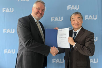 Zwei Personen im Anzug blicken freundlich in die Kamera, sie halten eine Urkunde in der Hand und geben sich die Hand.