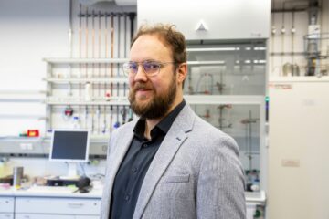 Eine Person mit Brille und Bart blickt lächelnd in die Kamera, im Hintergrund ist ein Computer-Bildschirm zu erkennen, an der Wand sind Leitungen befestigt.