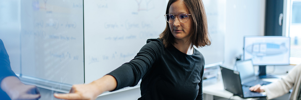 Eine Frau mit Brille und dunklen Haaren steht vor einem Whitboard. Sie deutet mit ihrem Zeigefinger auf etwas Geschriebenes auf dem Board. Im Hintergrund sieht man ein weiteres Whiteboard und zwei offene Laptops auf einem Tisch.