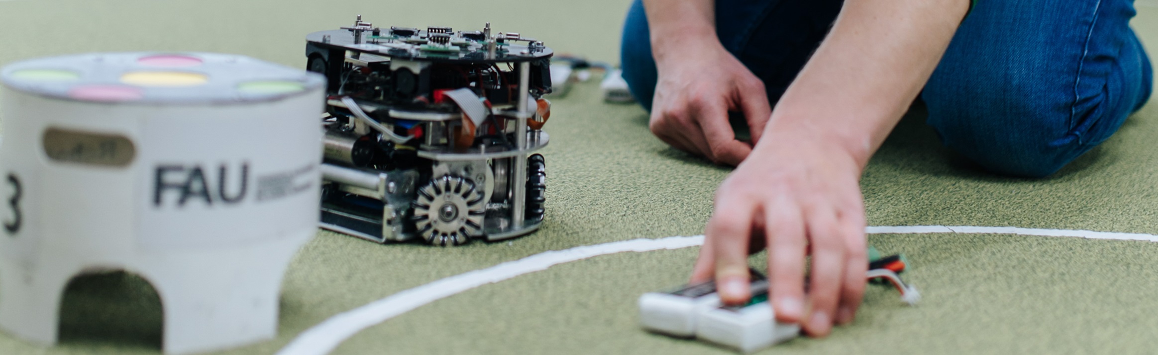 Auf der linken Bildseite sind zwei kleine Roboter zu sehen. Rechts daneben erkennt man die Hände eines Menschen außerhalb des Fotos.