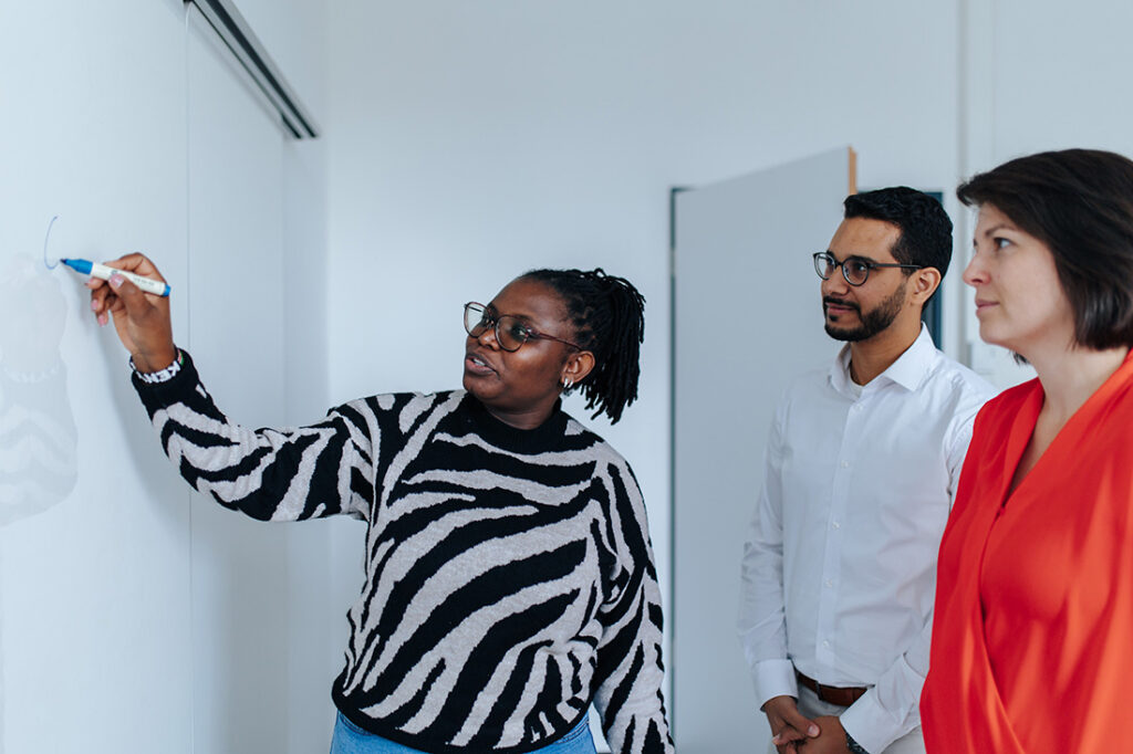 Eine Person steht vor einem Whiteboard, sie schreibt mit einem blauen Stift auf das Board. Zwei weitere Personen schauen auf das Whitboard.