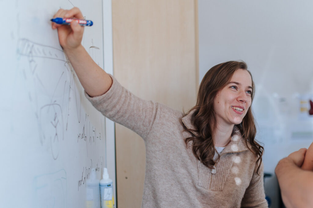 Eine junge Frau steht an einem Whiteboard und schreibt etwas an die Tafel. Sie erklärt etwas einer Person, die außerhalb des Bildes steht.