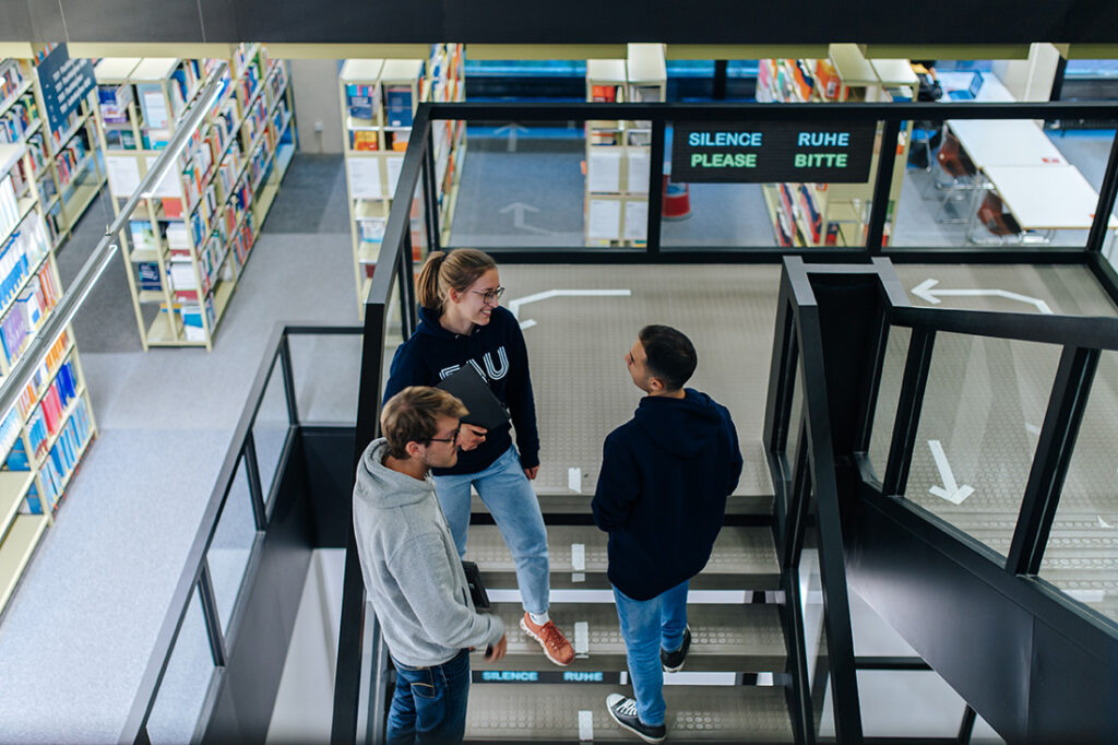 Drei Personen stehen auf einer Treppe beieinander und unterhalten sich. Um sie herum stehen Regale mit Büchern.