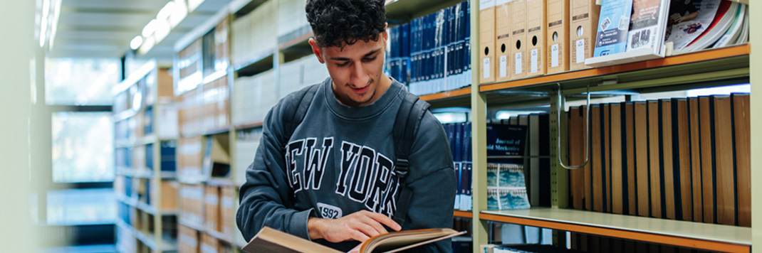 Ein junger Mann steht in einer Bibliothek zwischen den Bücherregalen und liest ein Buch.