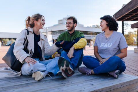 Eine Gruppe von drei jungen Menschen sitzt auf einer Holzbank auf einem Platz vor einem Gebäude.
