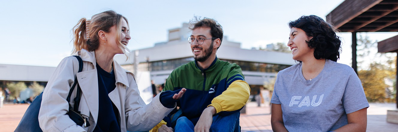 Zwei Frauen und ein Mann sitzen auf einer Holzbank auf dem Campus einer Universität.