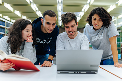 Eine Gruppe von vier jungen Menschen, zwei Männer und zwei Frauen, befinden sich in einer Bibliothek und blicken gemeinsam in einen Laptop.