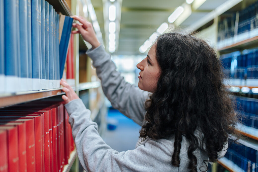 Eine junge Frau mit dunklen, langen Haaren greift nach einem Buch, das neben vielen anderen Büchern in einem Regal steht.