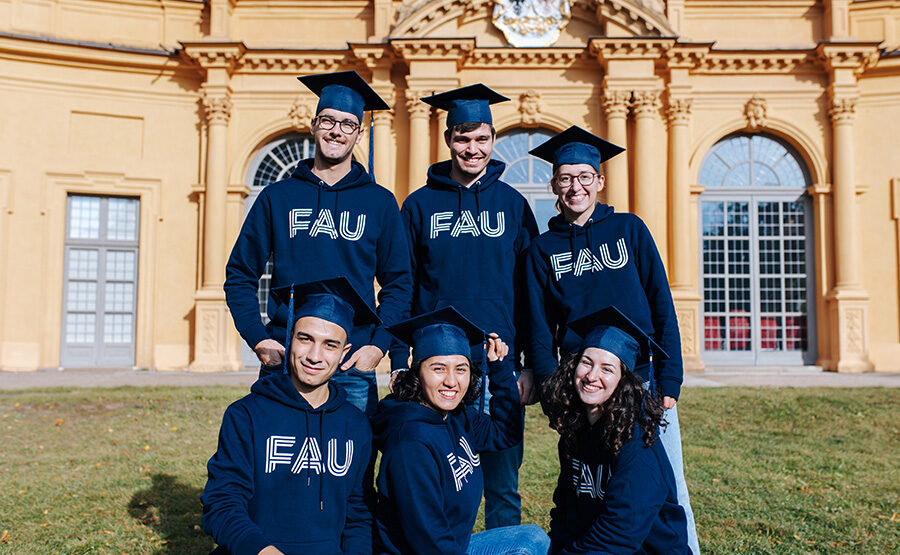 Eine Gruppe von sechs Personen steht vor einem orangenen Gebäude mit großen Fenstern. Alle tragen blaue Absolventenhüte und einen blauen Kapuzenpullover mit dem Aufdruck FAU.