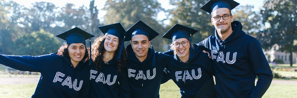 Eine Gruppe von fünf Personen steht in einem Park. Alle tragen Absolventenhüte einer Universität und liegen sich in den Armen.