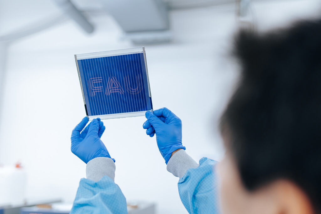 Eine Person mit blauen Handschuhen hält ein dunkelblaues Schild nach oben. Auf dem Schild steht aufgedruckt FAU.