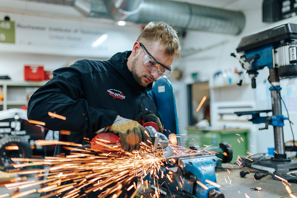 Ein Mann mit Schutzbrille steht in einer Werkstatt. Er schneidet mit einer Flex Metall, dabei fliegen Funken.