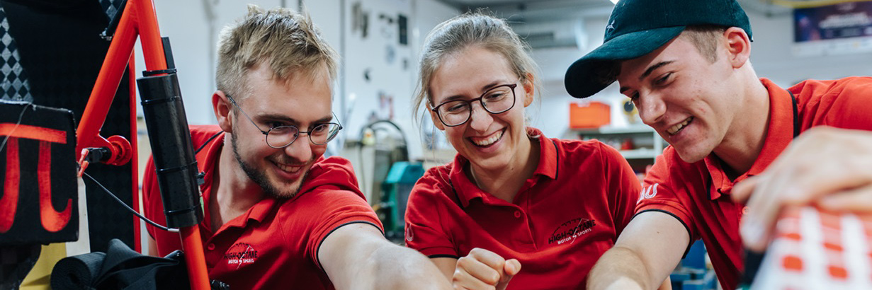 Eine Gruppe von zwei jungen Männern und einer jungen Frau arbeiten an einem Projekt in einer Werkstatt. Sie tragen rote T-Shirts. Der Mann auf der linken Seite deutet auf das Auto, an dem sie gerade arbeiten.