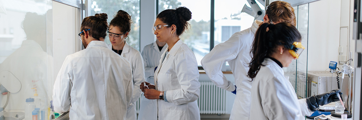 Mehrere Personen mit Schutzbrillen und weißen Mänteln stehen in einem Labor, sie sprechen und arbeiten miteinander.