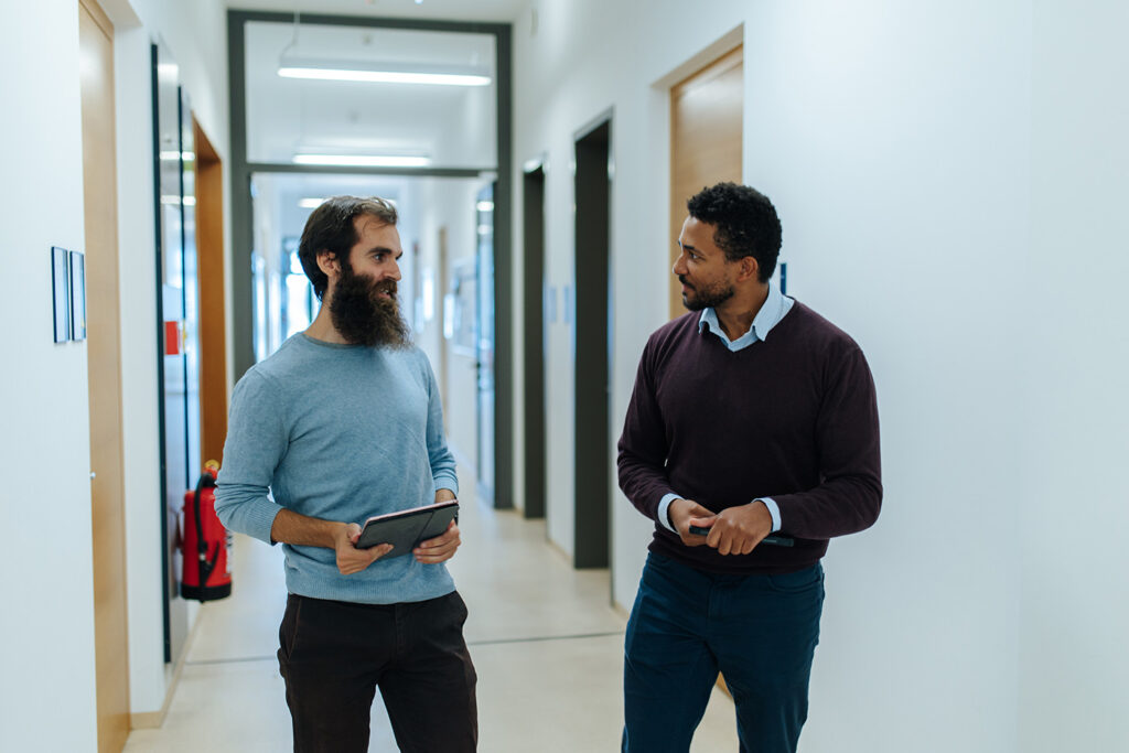 Zwei Männer mit dunklen Haaren, einer davon mit Bart, stehen in einem Gebäudeflur mit vielen Türen auf beiden Seiten. Die beiden Personen blicken sich an und sprechen miteinander.