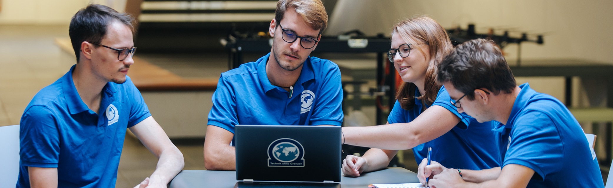 Drei Studierende, drei Männer und eine Frau, tragen blaue T-Shirts, sitzen an einem Tisch und blicken gemeinsam auf einen Laptop.