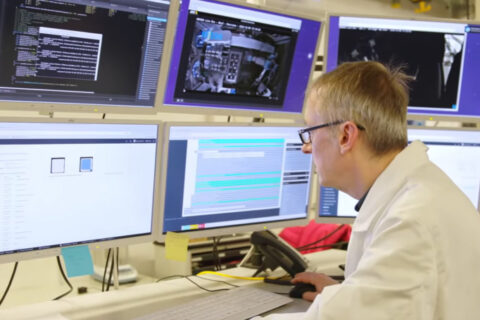 Eine Person in weißem Laboranzug sitzt vor 6 Bildschirmen auf denen in verschiedenen Farben markierte Texte zu sehen sind. In der Hand hält er eine PC-Maus.