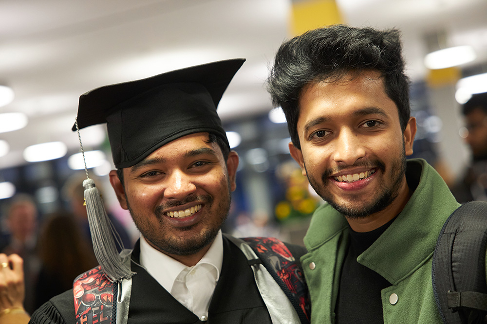 2 Personen mit schwarzen Haaren blicken freundlich lächelnd in die Kamera. Eine Person trägt einen schwarzen Absolventenhut.