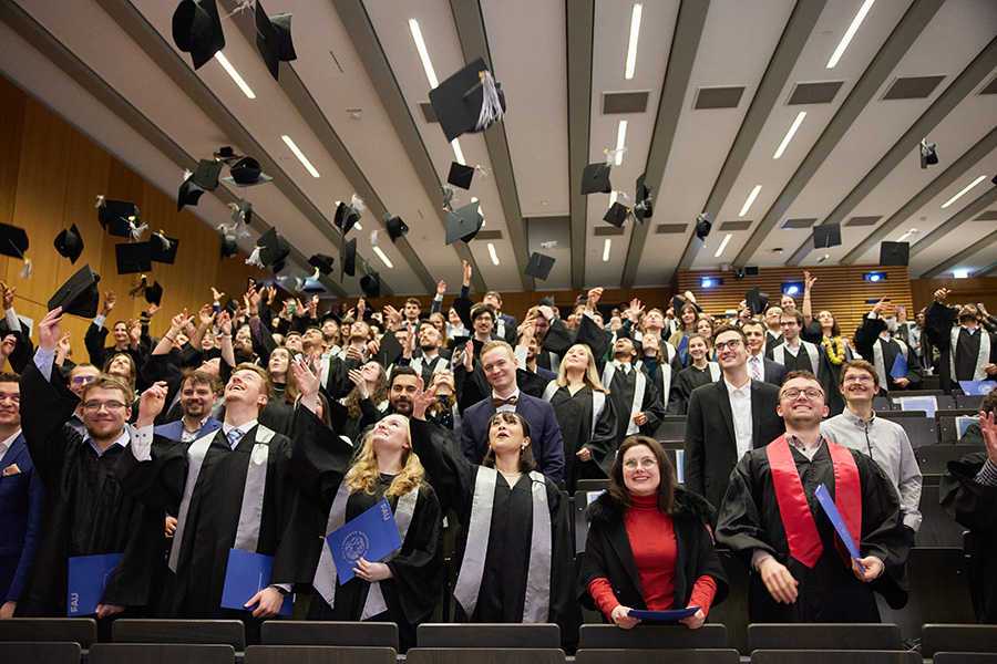 Viele lachende Personen stehen in einem Hörsaal und werfen ihre Absolventenhüte in die Höhe.