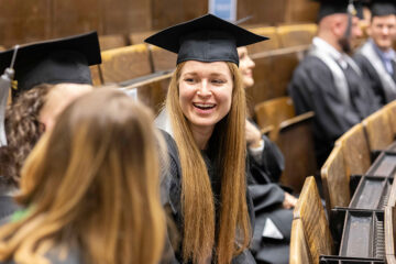 Mehrere Personen sitzen in einer Reihe von braunen Klappstühlen. Die Personen tragen schwarze Absolventenhüte.