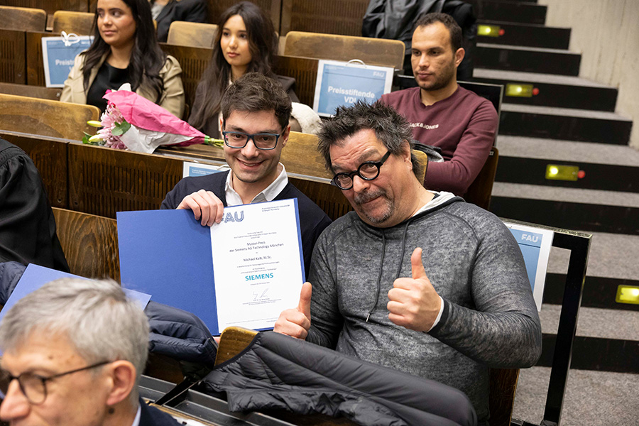 In Sitzreihen in einem Hörsaal sitzen mehrere Personen. Zwei Personen schauen direkt in die Fotoakamera und lächeln. Die eine Person hält den Daumen in die Luft, die andere Person zeigt eine offene blaue Mappe mit einem weißen Papier darin.