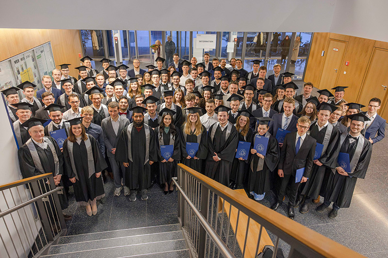 Am Fuße einer Treppe stehen viele Personen nebeneinander und blicken lachend in die Kamera. Viele tragen einen schwarzen Talar und einen schwarzen Absolventenhut. In ihren Händen halten sie eine blaue Mappe. Im Hintergrund ist eine Glaswand zu erkennen.