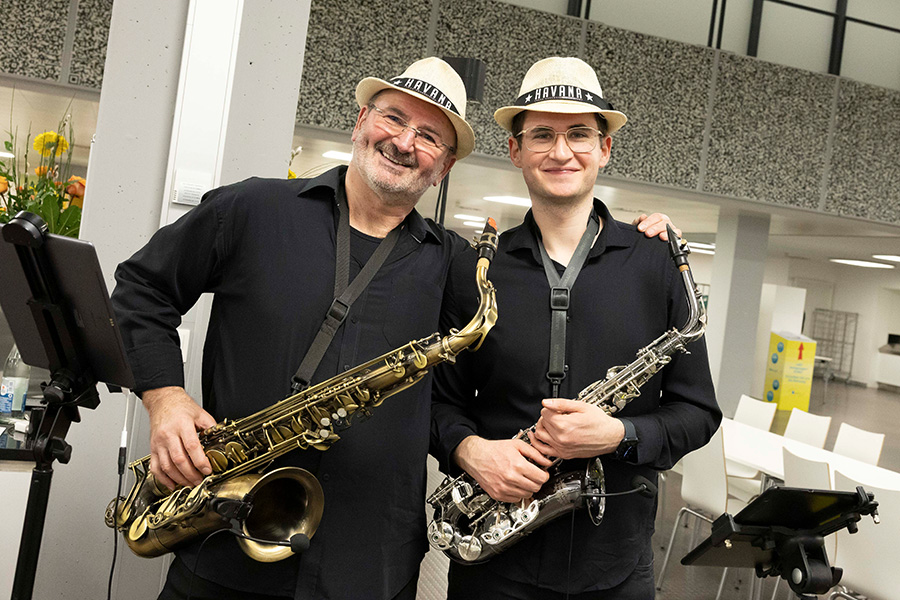 Zwei Personen mit Brille und weißen Hüten auf dem Kopf halten in ihrer Hand ein Saxophon und lachen in die Kamera.
