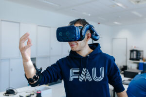 Eine Person trägt eine blaue Virtuell-Reality-Brille und blickt damit auf die eigene Hand.
