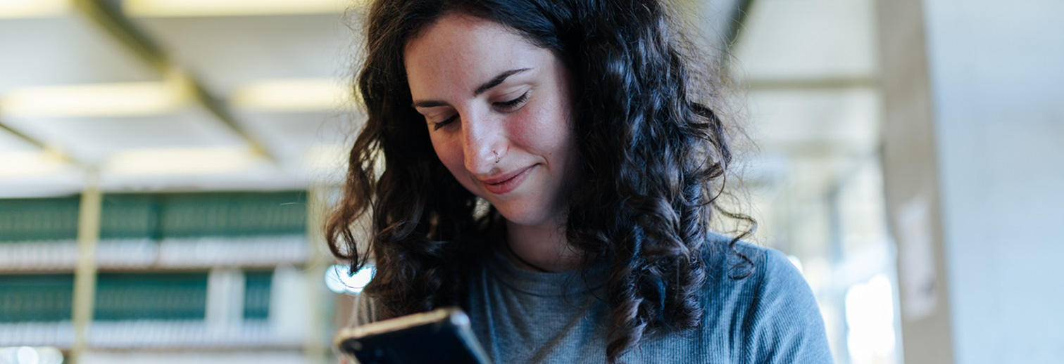 Eine junge Frau mit dunklen lockigen Haaren steht in einer Bibliothek und blickt auf ein Handy.