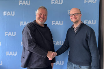Zwei Personen sehen lächelnd in die Kamera und geben sich dabei die Hand. Im Hintergrund ist eine hellblaue Wand mit dem Logo FAU.