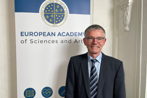 Eine Person im blauen Anzug, hellem Hemd und gestreifter Krawatte steht vor einem RollUp mit dem Logo der European Academy of Sciences and Art.